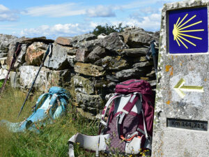 Una señalización del Camino de Santiago, junto dos mochilas de peregrinos apoyadas en un pequeño muro de piedra